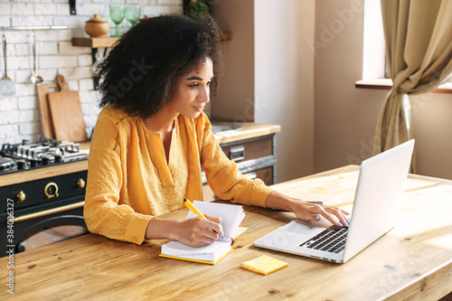 An african-american young woman is using laptop computer for remote work or studying online, she takes notes watching webinars or classes at kitchen table at home photo