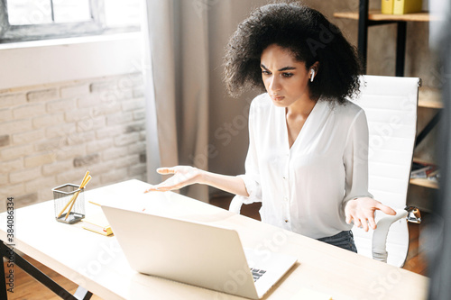 A puzzled african woman looks at laptop screen and spreads her hands in disbelief. Misunderstanding at work