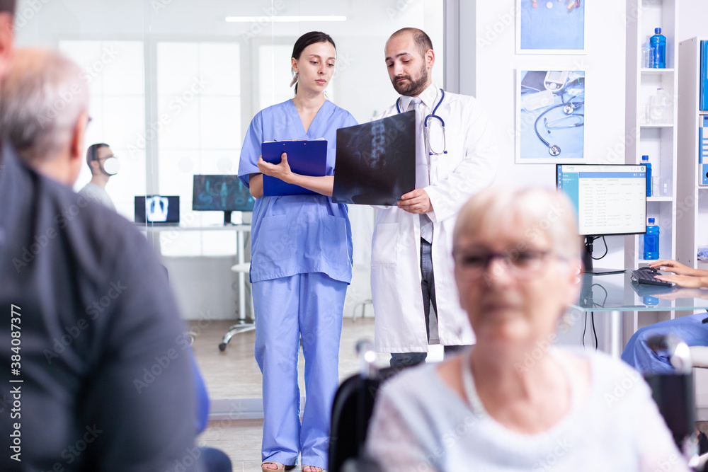Nurse looking at radiography while talking with doctor about patient diagnosis in hospital waiting area. Invalid old woman in wheelchair. Senior man waiting for medical examination.