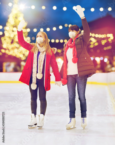 christmas, winter and leisure concept - two women or friends wearing face protective medical masks for protection from virus disease waving hands at outdoor skating rink over holiday lights background