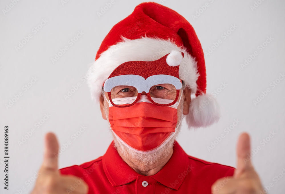 A senior man dressed in red as Santa Claus wearing a surgical mask due to the coronavirus and makes a sign of optimism. Merry christmas at the time of the covid-19 virus. A senior man dressed in red as Santa Claus wearing a surgical mask due to the coronavirus and makes a sign of optimism. Merry christmas at the time of the covid-19 virus.