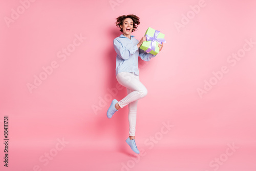 Full length profile photo of pretty funny lady jumping high up hold big large gift box birthday girl hold boyfriend surprise wear blue shirt pants shoes isolated pink color background