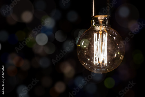 Glowing vintage old one tungsten lightbulb hanged on ceiling against black wall bokeh background. Retro styled decoration electric lamp. Christmas interior decor concept