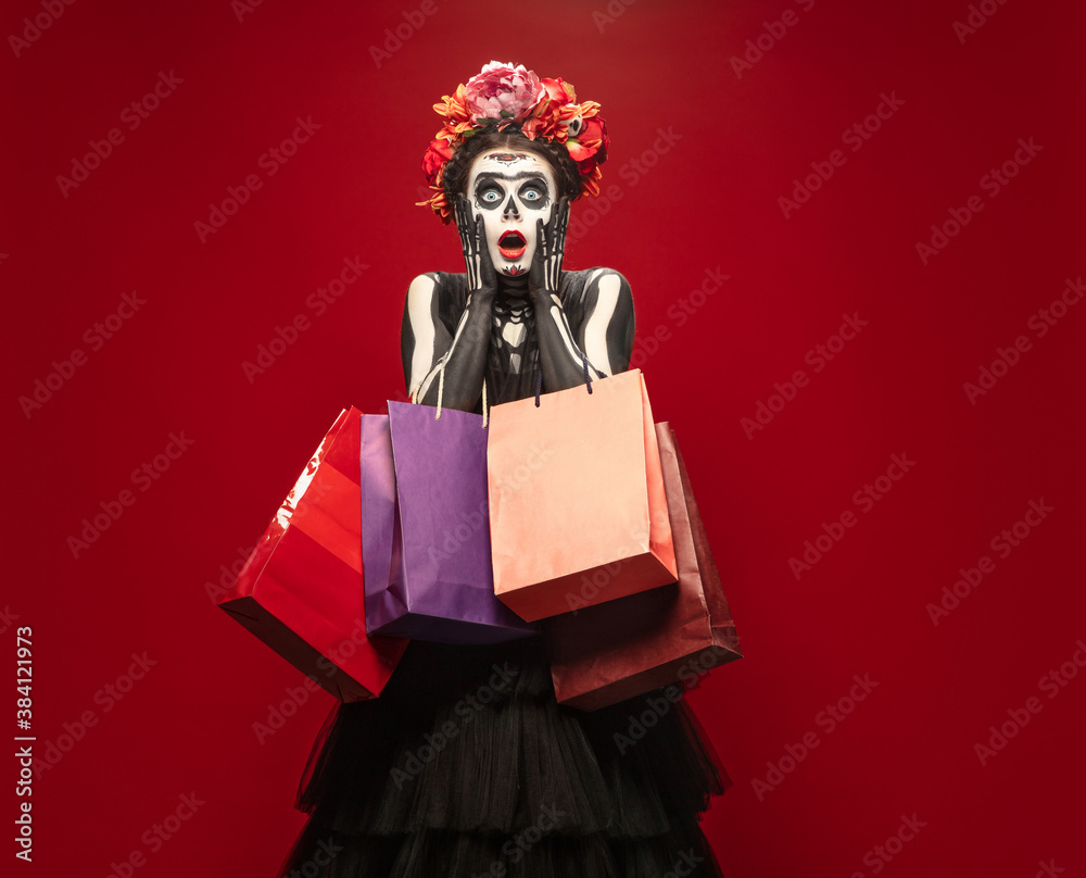 Shopping. Young girl like Santa Muerte Saint death or Sugar skull with bright make-up. Portrait isolated on red studio background with copyspace. Celebrating Halloween or Day of the dead. Black friday Shopping. Young girl like Santa Muerte Saint death or Sugar skull with bright make-up. Portrait isolated on red studio background with copyspace. Celebrating Halloween or Day of the dead. Black friday