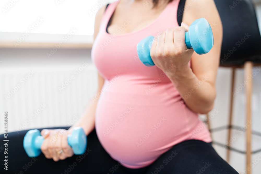 Beautiful pregnant woman with light blue dumbbells sitting on fitball and training in living room at home. Side view, body part, lifting dumbbells, active and sportive pregnancy concept. Beautiful pregnant woman with light blue dumbbells sitting on fitball and training in living room at home. Side view, body part, lifting dumbbells, active and sportive pregnancy concept.