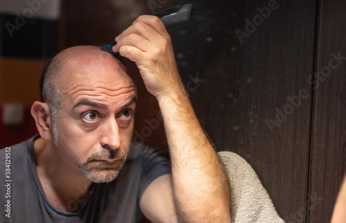 A middle-aged Caucasian man cuts his hair in the bathroom. While placing the clipper on your head check with your eyes in the mirror.