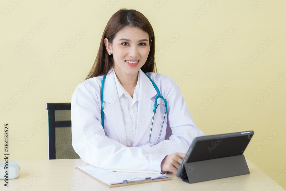 Asian beautiful young smiling female doctor sitting in office at hospital. On table has a clipboard and digital tablet computer.