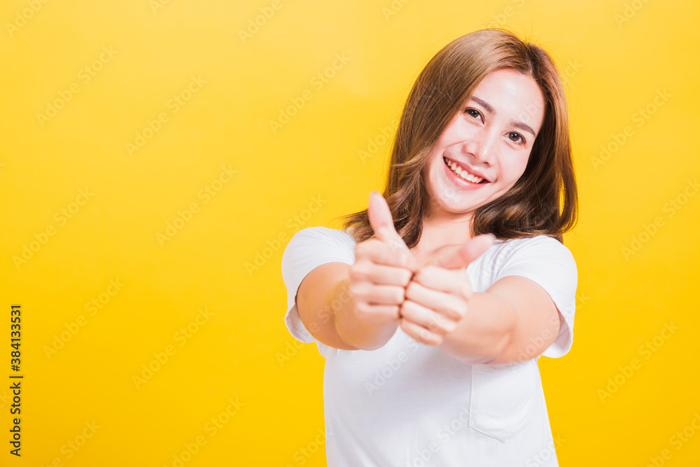 Portrait Asian Thai beautiful happy young woman smiling wear white t-shirt standing successful woman giving two thumbs up gesture sign in studio shot, isolated on yellow background with copy space Portrait Asian Thai beautiful happy young woman smiling wear white t-shirt standing successful woman giving two thumbs up gesture sign in studio shot, isolated on yellow background with copy space