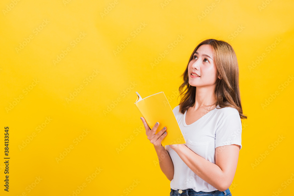 Portrait Asian Thai beautiful happy young lifestyle woman stands holding yellow book or diary she show thumb up finger and looking up away, studio shot isolated on yellow background, with copy space