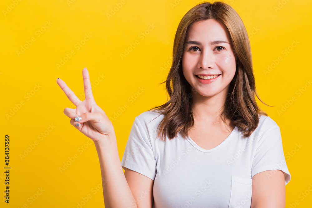 Asian Thai happy portrait beautiful cute young woman smile standing wear t-shirt showing finger making v-sign symbol near eye looking to camera, isolated studio shot yellow background with copy space Asian Thai happy portrait beautiful cute young woman smile standing wear t-shirt showing finger making v-sign symbol near eye looking to camera, isolated studio shot yellow background with copy space