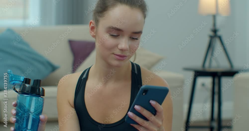 Sporty woman drinking water and use phone while resting after training at home. Healthy