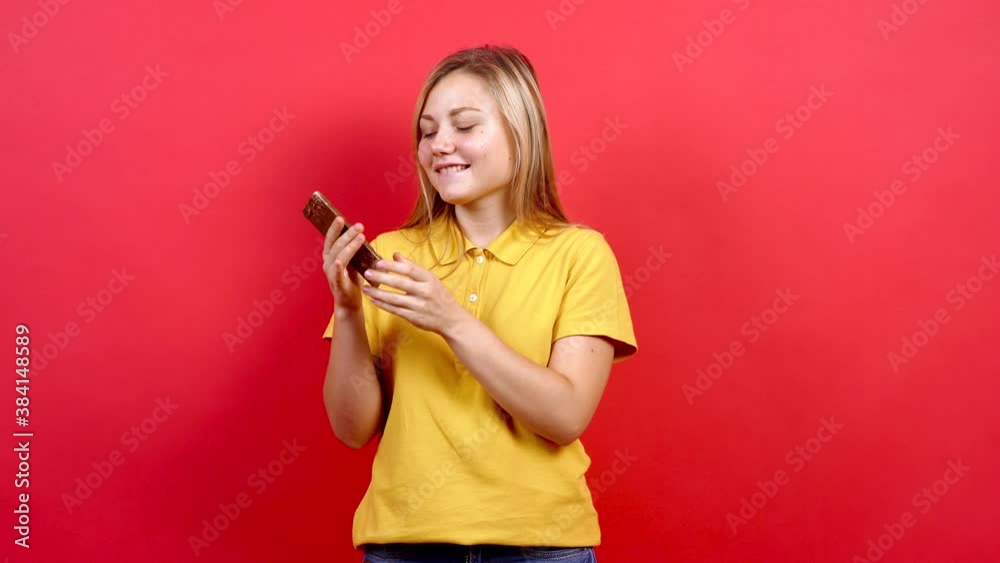 Cute and a little fat girl in a yellow T-shirt holds a chocolate in her hand. She is excited and likes sweets. Concept of unhealthy nutrition. Isolated on a red background.