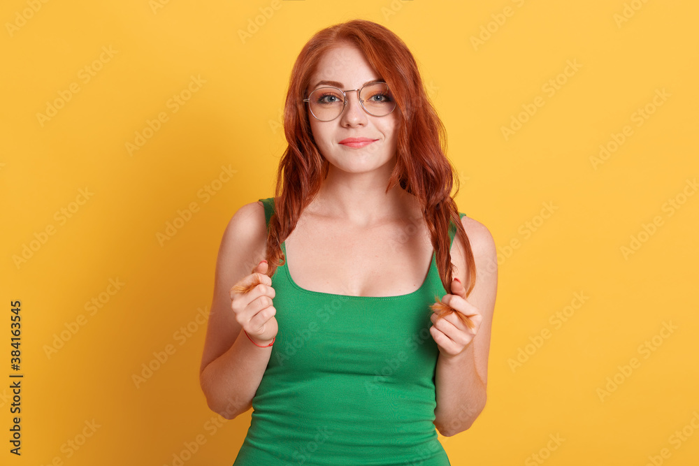Redhead girl isolated over yellow background with charming smile, wearing glasses and green t shirt, keeping hands on her curls, lady with pleasant appearance expressing positive emotions. Redhead girl isolated over yellow background with charming smile, wearing glasses and green t shirt, keeping hands on her curls, lady with pleasant appearance expressing positive emotions.