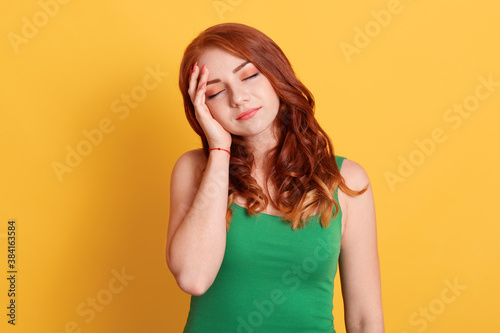 Young good looking red haired female stands with closed eyes isolated over yellow background, keeps hands on temples, suffering from terrible headache, girl wears green t shirt.