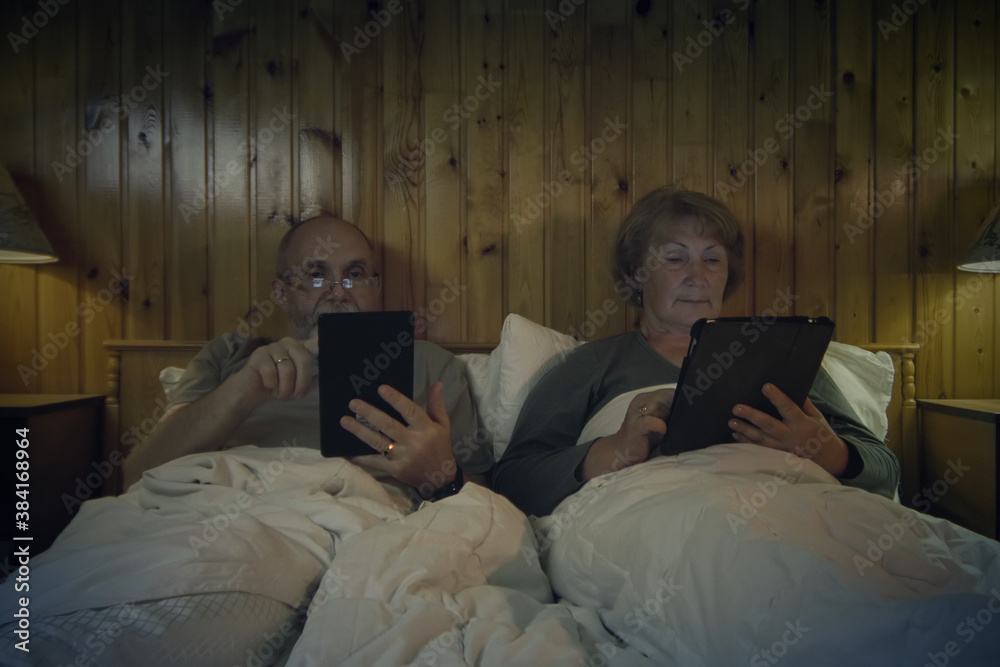 Senior Couple Wearing Pajamas Lying In Bed Reading On Digital Tablet. Senior Couple In Bed Before Go To Sleep