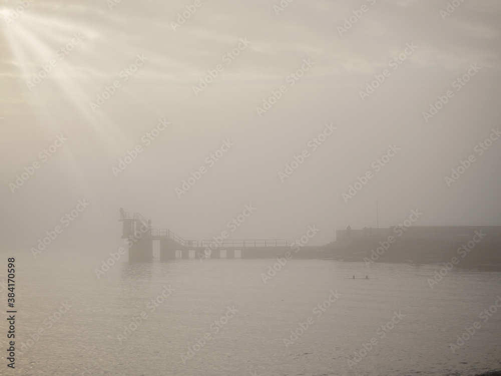 Silhouette of Blackrock public diving tower in a fog. Cloudy sky over ocean with sun beams bursting through clouds. Calm and tranquil atmosphere. Salthill, Galway city, Ireland