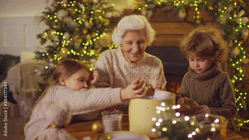 Medium closeup of lady putting Christmas homemade gifts in box with craft paper with grandchildren at sides in decorated room Medium closeup of lady putting Christmas homemade gifts in box with craft paper with grandchildren at sides in decorated room