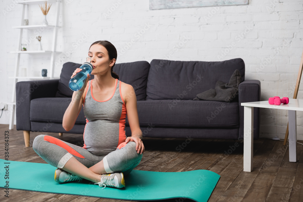 pregnant woman sitting on fitness mat while drinking water from sports bottle - obrazy, fototapety, plakaty 