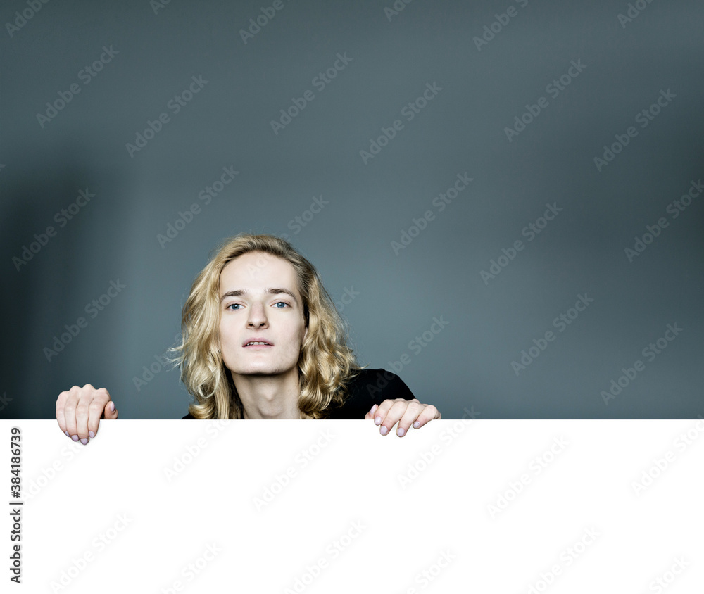A young attractive man peeks out from above a white wall on a gray background. Looks surprised laid his head in his hands. The elbows are apart.