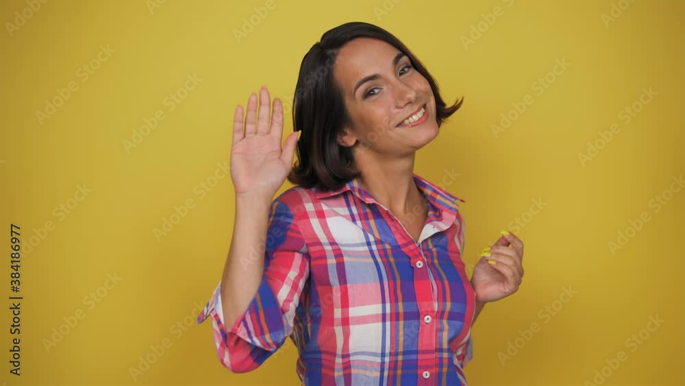 Young woman with brown hair happily smile in the camera. Woman standing in the yellow background. Woman wave to the camera with her hand. High quality 4k footage Young woman with brown hair happily smile in the camera. Woman standing in the yellow background. Woman wave to the camera with her hand. High quality 4k footage