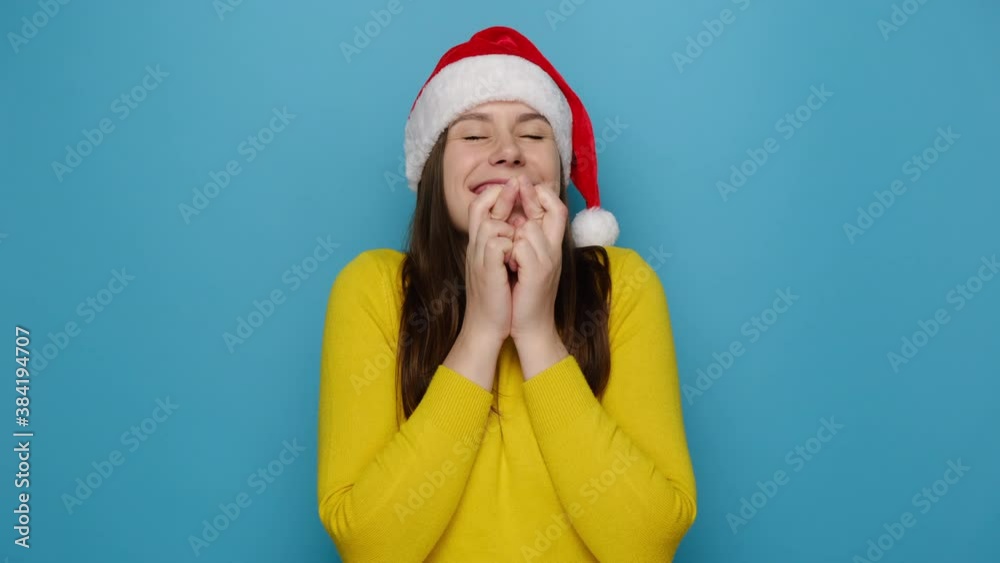 Making Christmas wish concept. Happy cute young woman stands with fingers crossed, awaits miracle happen, wears red hat and yellow sweater, believes in good luck, isolated on blue studio background
