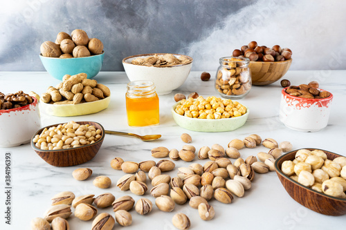 Various nuts (hazelnuts, almonds, walnuts, cashews, pine nuts) peeled and in shell in wooden and ceramic bowls are on a white table. In the center there is a small jar of honey and scatterer pistachio photo