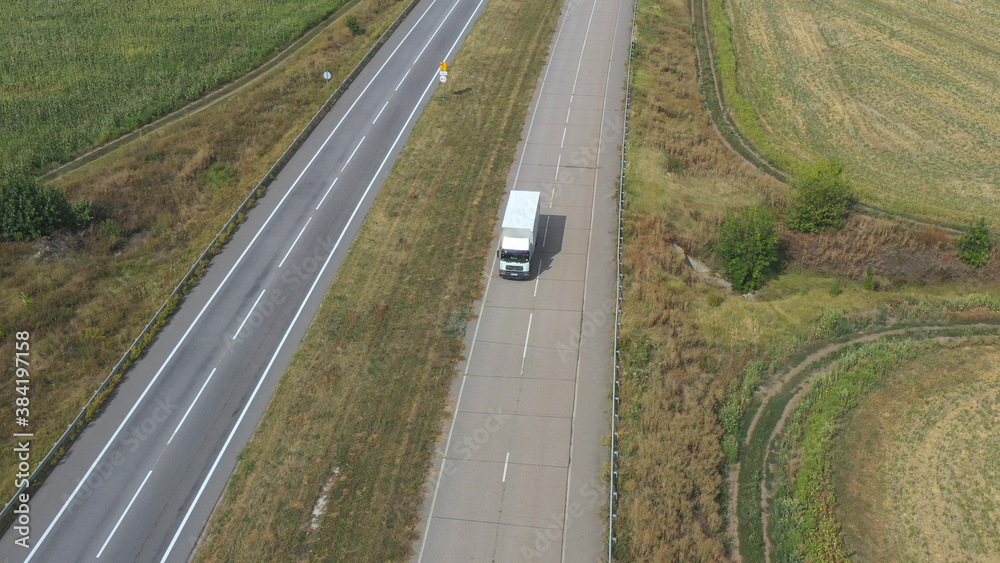 Aerial view of truck with cargo trailer driving on empty road and transporting goods. Flying over delivery lorry moving along highway in countryside. Beautiful rural environment around. Slow motion