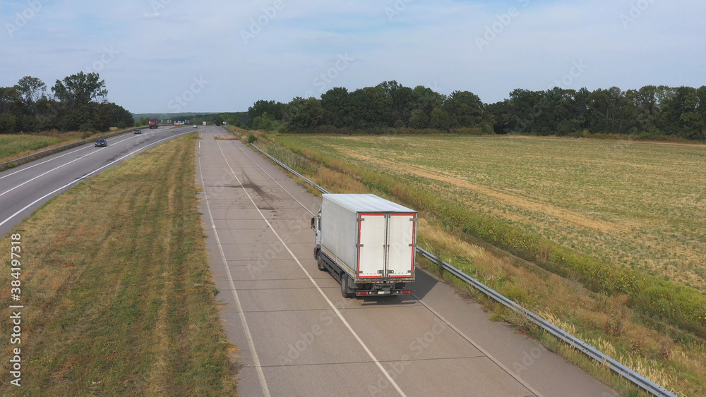 Aerial shot of truck with cargo trailer driving on road and transporting goods. Flying over delivery lorry moving along highway passing in countryside with scenic nature environment around. Back view