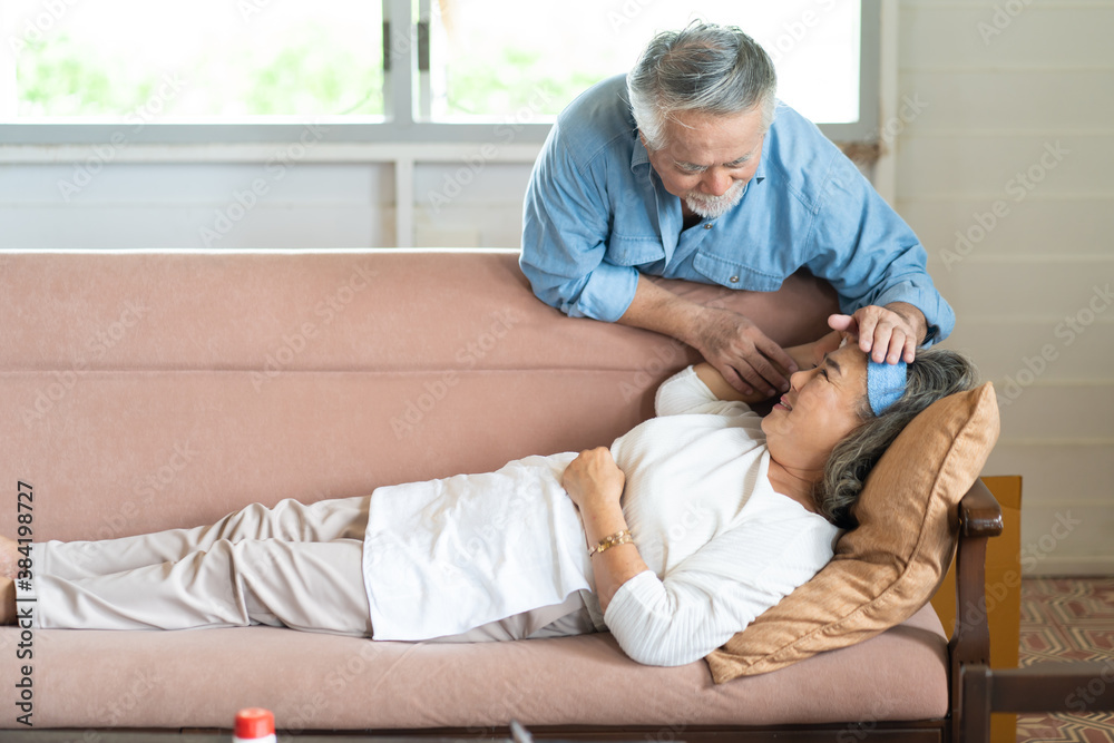 Elderly Asian man checking fever temperature of senior wife lying on sofa at home. Elderly Asian man checking fever temperature of senior wife lying on sofa at home.