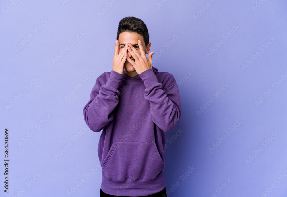Young caucasian man isolated on purple background blink at the camera through fingers, embarrassed covering face. Young caucasian man isolated on purple background blink at the camera through fingers, embarrassed covering face.
