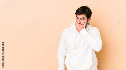 Young man isolated on beige background thoughtful looking to a copy space covering mouth with hand.