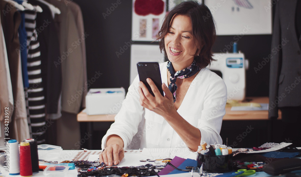 Fashion designer with beaming smile sitting at table in tailor workshop and browsing smartphone