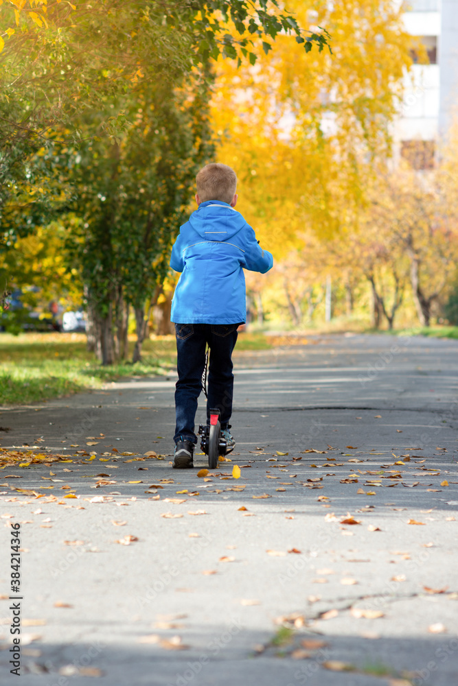 A boy in a blue jacket rides a scooter down a city street in autumn A boy in a blue jacket rides a scooter down a city street in autumn