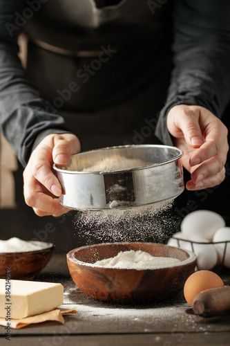 Chef hands pouring flour powder on raw dough using sieve on a black background, Cooking process.