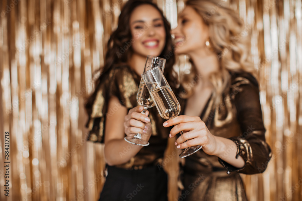 Girls in bronze-colored dresses clink glasses with champagne on gold background - obrazy, fototapety, plakaty 