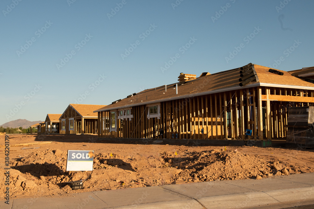 A new house construction with the wood frame before the siding  stands next to an empty dirt lot with a sold sign, horizontal view.