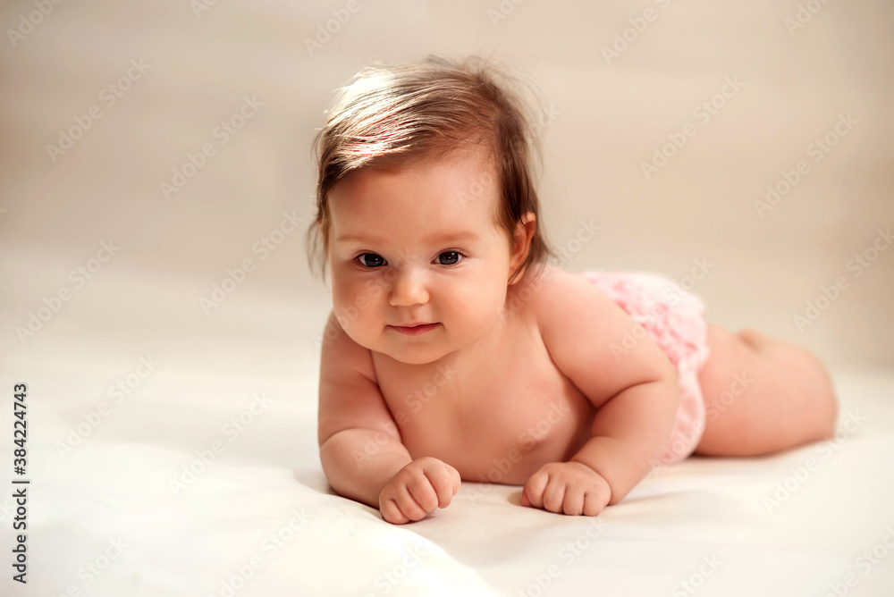 Smiling infant baby girl in pink pants lying on belly on light yellow (champagne colored) background with sun fleck on her head. Early intellectual and cognitive development concept Smiling infant baby girl in pink pants lying on belly on light yellow (champagne colored) background with sun fleck on her head. Early intellectual and cognitive development concept