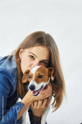 A portrait of a young caucasian woman in a denim jacket with her pet, jack russell terrier. She is slightly hiding behind the happy dog, kissing him and looking into the camera. White background.