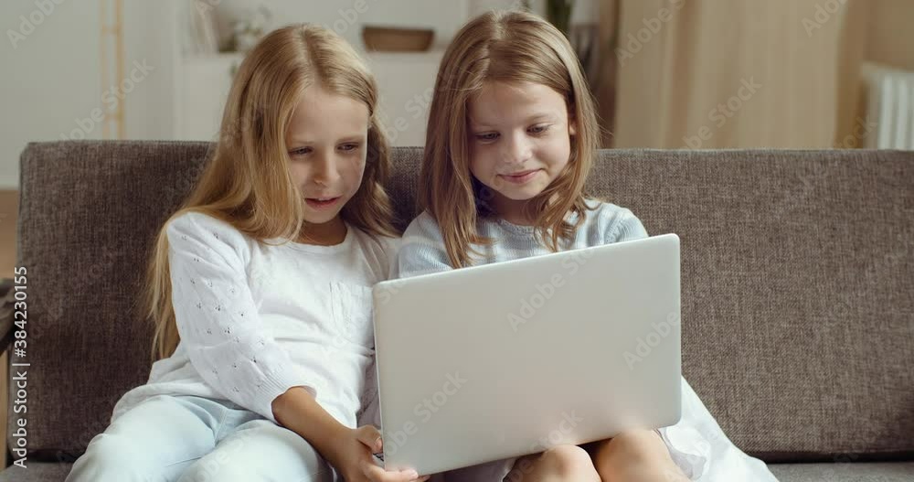 Two little girls sitting on couch looking at laptop, blonde sisters at home together watching movie or video online in computer screen, couple of girls friends spend time together laughing having fun Two little girls sitting on couch looking at laptop, blonde sisters at home together watching movie or video online in computer screen, couple of girls friends spend time together laughing having fun