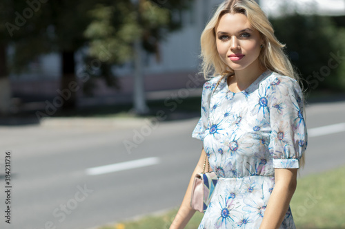 A young girl in a light blue dress walks embarrassedly down the street on a sunny summer day against the backdrop of a blurred background photo