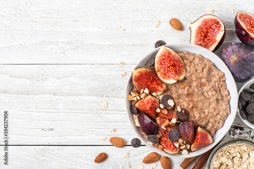 Bowl of oatmeal porridge with figs, cinnamon, chocolate and almonds on white wooden background. top view