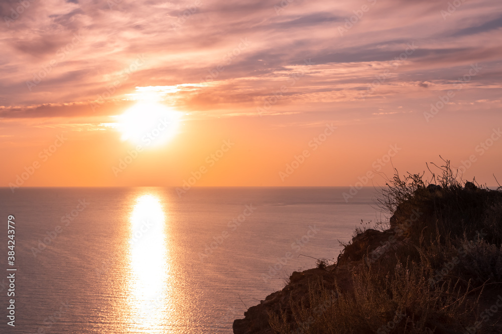 A large crimson Sun sets over the horizon against a cloudy sunset pink sky. Twilight sun shines on the top of the cliff. The edge of the cliff turns into a abyss. Black evening sea below the cliffs.