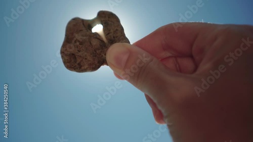 Hand holding small stone with a hole sun is shining through. Light leaks and flares. Summer beach memories concept