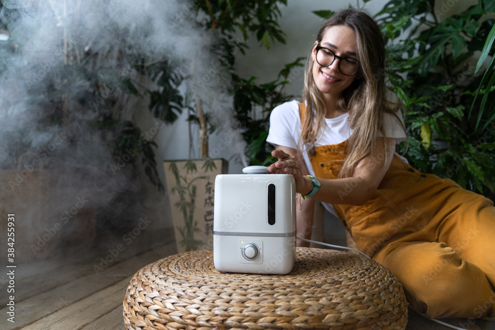 Woman gardener in overalls feeling fresh, sitting on the wooden floor, using air humidifier in home garden during heating period, houseplants on background. Plant care.  - obrazy, fototapety, plakaty 