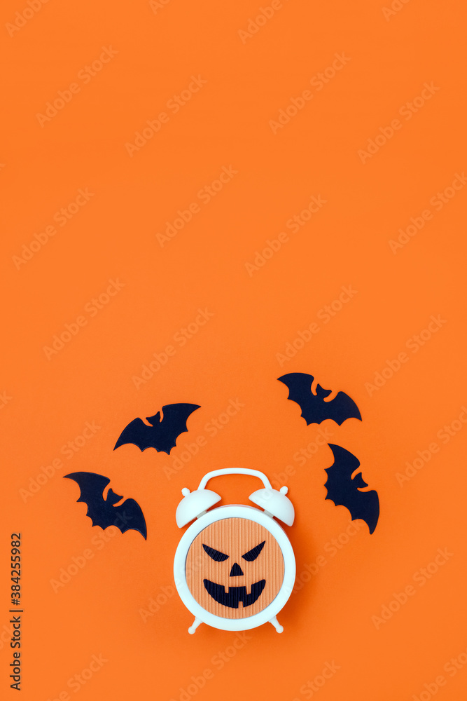 Alarm clock with evil face and fly bats made of orange and black cardboard. Halloween minimal concept. Vertical postcard
