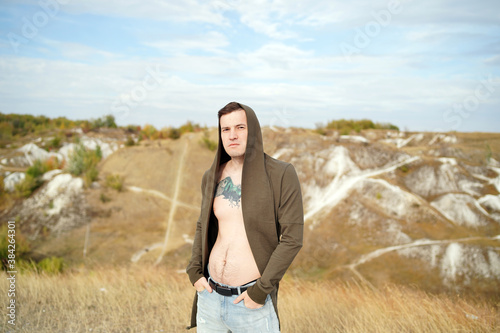 Tattooed man with bare torso in cape and jeans stands in hilly terrain. Adult male poses on cloudy day in countryside.