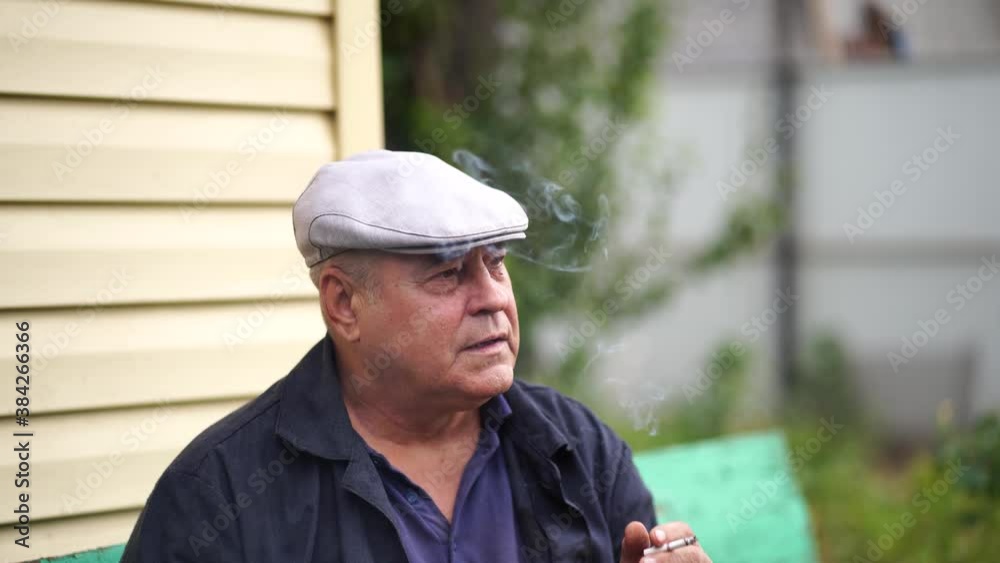 Close up of mature man smokes, sitting on bench in garden. Older male resting and smoking cigarette outdoors.