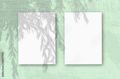 2 vertical sheets of textured white paper on soft green table background. Mockup overlay with the plant shadows. Natural light casts shadows from an willow branch. Horizontal orientation