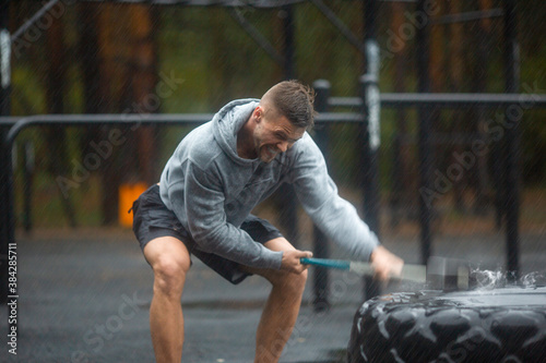 Heavy outdoor training on a rainy day - a man hits a tire with a sledgehammer.