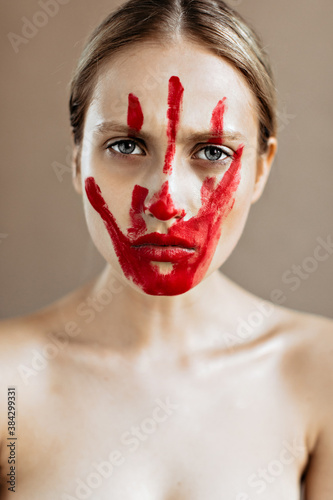 Gray-eyed blond woman with bloody hand print on her face looks sternly at camera